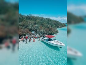 A lively group enjoys a beach day in shallow turquoise waters, with a boat anchored nearby, perfect for a fun-filled outing.