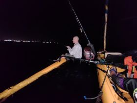 A fisherman enjoys a night fishing experience from a kayak, with city lights twinkling in the background — perfect for adventurous anglers seeking a unique outing.