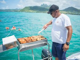 A chef grills delicious food on a boat while guests enjoy swimming in the vibrant turquoise waters — a perfect blend of culinary delight and aquatic fun.