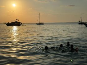 A group of swimmers enjoys a beautiful sunset in the water, with boats anchored nearby, creating a perfect backdrop for a memorable beach experience.