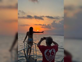 A woman poses at the bow of a boat during a stunning sunset while a man captures the moment on his phone — a perfect scene for romantic boat trips or sunset cruises.