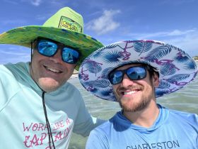 Two friends enjoy a sunny day at the beach, sporting vibrant hats and sunglasses — perfect for a fun day on the water.