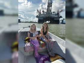 Two girls dressed in colorful mermaid tails enjoy a boat ride with an oil rig in the background, capturing a whimsical moment on the water.
