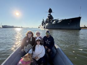 A cheerful group poses on a small boat with a historic battleship in the background, enjoying a sunny day on the water.