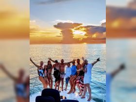 A lively group celebrates at sunset aboard a yacht, showcasing joy and camaraderie against a stunning backdrop of colorful skies and calm waters.