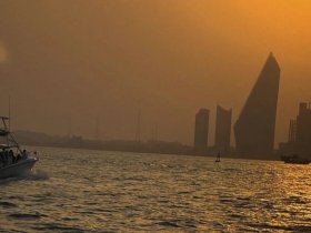 A picturesque sunset view featuring a boat cruising past a stunning city skyline, with a paddleboarder enjoying the tranquil waters — a perfect scene for evening adventures.