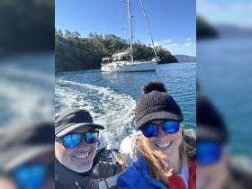 A cheerful couple takes a selfie while enjoying a sailing adventure, with a beautiful backdrop of a sailboat and serene waters.