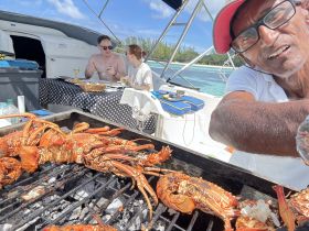 A chef prepares fresh grilled lobster on board, while guests enjoy a meal in the background — a perfect culinary experience on the water.
