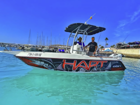 Two men stand on the deck of a fishing boat named 'HART' in clear turquoise waters, showcasing a vibrant day on the water.