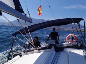 Two crew members navigate a sailboat through the ocean, with stunning mountain views in the background — a perfect representation of a sailing adventure.