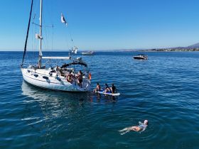A lively scene featuring a group enjoying water activities around a yacht, with paddleboarding and swimming in clear blue waters — perfect for summer adventures.