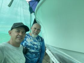 Two individuals pose for a photo in a covered area, with a boat partially visible in the background.