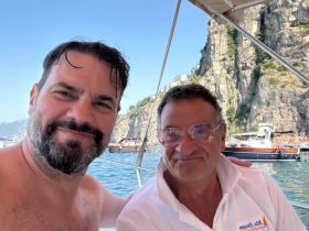Two men enjoy a cheerful moment aboard a boat with stunning coastal scenery in the background — a perfect snapshot of a memorable boating experience.