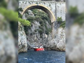 A striking red boat navigates through a narrow passage beneath a historic archway, showcasing the stunning coastal scenery.