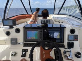 A view of the helm inside a boat, showcasing the navigation equipment and controls, with two individuals visible in the background enjoying the ride.