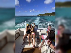 A lively group enjoys a fun day on the water, taking selfies and sipping drinks aboard a boat, surrounded by stunning turquoise waters and a beautiful coastline.