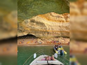 Guests relax on the deck of a boat while exploring a stunning cave beach, with kayakers enjoying the clear waters — a perfect day for adventure and relaxation.