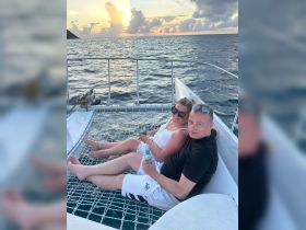 A couple enjoys drinks while relaxing on the netting of a catamaran at sunset, creating a perfect backdrop for romantic moments on the water.