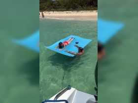 A child enjoys a sunny day on a floating mat while an adult assists in the clear waters near a sandy beach, capturing the essence of summer fun and relaxation.