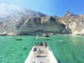 A group enjoys a refreshing swim in crystal-clear waters near a stunning rocky coastline, with a boat anchored nearby — perfect for a day of adventure and relaxation.