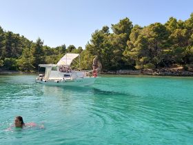 A group enjoys a sunny day on the water, with one person swimming and others relaxing on the boat amidst stunning turquoise waters and lush greenery.