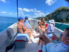 A lively group enjoys a sunny day on the water, relaxing on a boat with drinks in hand, surrounded by stunning ocean views and cliffs.