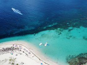 A stunning aerial view of a beach with colorful umbrellas and people enjoying the clear waters, complemented by boats anchored nearby — a perfect tropical getaway.