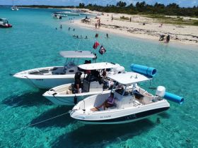 A vibrant scene at a sandbar with multiple boats anchored, where guests enjoy swimming and relaxing on the beach — a perfect day for water activities and fun in the sun.