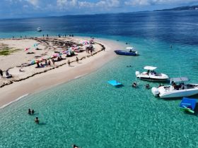 A vibrant scene at a sandbar with groups enjoying the sun and swimming in crystal-clear waters, surrounded by boats and colorful umbrellas — a perfect day for beachgoers and water enthusiasts.