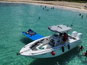 A lively scene featuring a boat anchored in clear waters, with guests enjoying swimming and relaxing on a floating mat — perfect for a fun day at the beach.