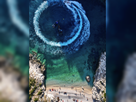 Aerial view of boats creating waves in clear turquoise waters, with beachgoers enjoying the sun and sand below — a perfect scene of summer fun and water activities.