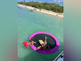 Two girls relax on a floating mat in crystal-clear waters, enjoying snacks and drinks while surrounded by beachgoers — a perfect day for fun in the sun.