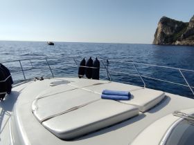 A serene view of a yacht's deck with plush seating, set against a backdrop of calm blue waters and distant cliffs, perfect for relaxation and enjoying the sea.
