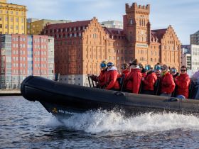 A group of adventurers in red jackets enjoy a thrilling ride on a RIB boat, with a stunning cityscape backdrop featuring historic architecture.