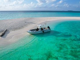 Aerial view of a speedboat anchored on a pristine sandbar, surrounded by crystal-clear turquoise waters — an ideal spot for fishing or relaxing in the sun.