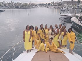 A vibrant group of friends poses on the deck of a yacht, dressed in matching yellow outfits, celebrating together in a picturesque marina setting.