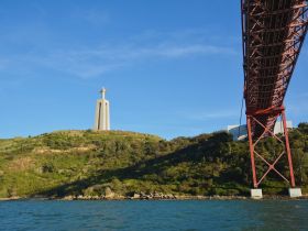 A stunning view of the iconic Christ the King statue overlooking the waterfront, framed by the impressive red bridge — a perfect representation of Lisbon's scenic beauty.