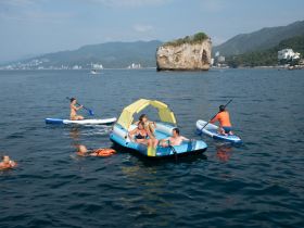 A lively group enjoys a day on the water, paddleboarding and swimming near a scenic rock formation — perfect for summer adventures and beach outings.