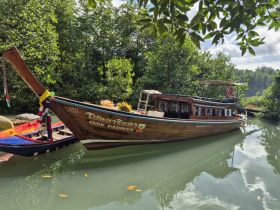 A beautifully crafted wooden boat named 'Chok Narinda 2' is docked amidst lush greenery, showcasing traditional design and serene waters, perfect for exploring mangroves.