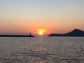 A stunning sunset over the harbor, featuring a lighthouse silhouette and distant mountains — a picturesque moment perfect for evening boat cruises.