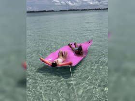 Two friends relax on a vibrant pink floating mat in crystal-clear waters, enjoying a perfect day at the beach.