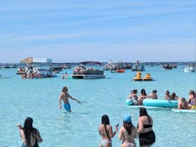 A vibrant scene at a sandbar with groups enjoying the water, swimming, and relaxing on inflatable mats, surrounded by various boats — a perfect summer getaway.