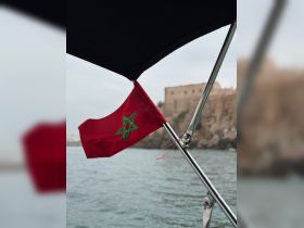 A close-up view of the Moroccan flag waving from a boat, with a historic fort in the background, showcasing the maritime culture of Morocco.