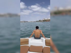 A young person enjoys a moment of relaxation on the edge of a boat, overlooking a scenic waterfront with buildings and boats in the background.