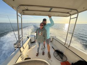 A couple enjoys a scenic moment on a fishing boat, with the sun setting over the ocean — a perfect backdrop for a memorable day on the water.