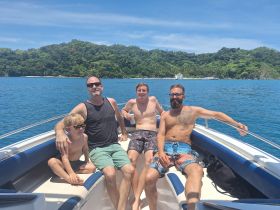 A joyful group of four, including a child, relaxes on a boat under the sun, enjoying a beautiful day on the water surrounded by lush greenery.