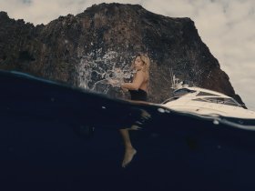 A woman enjoys a refreshing swim, creating splashes in the water, with a luxurious boat and a stunning rocky coastline in the background — a perfect scene for adventure seekers.