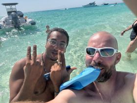 Two friends enjoy a fun day at the beach, posing for a selfie with a boat in the background — perfect for capturing memorable moments on the water.