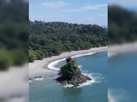 A stunning aerial view of a secluded beach with lush greenery and a rocky outcrop, showcasing the natural beauty of the coastline.