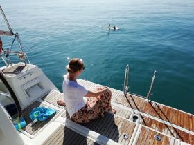 A woman relaxes on the deck of a boat while two individuals swim nearby in calm waters, capturing a serene moment of leisure on the water.